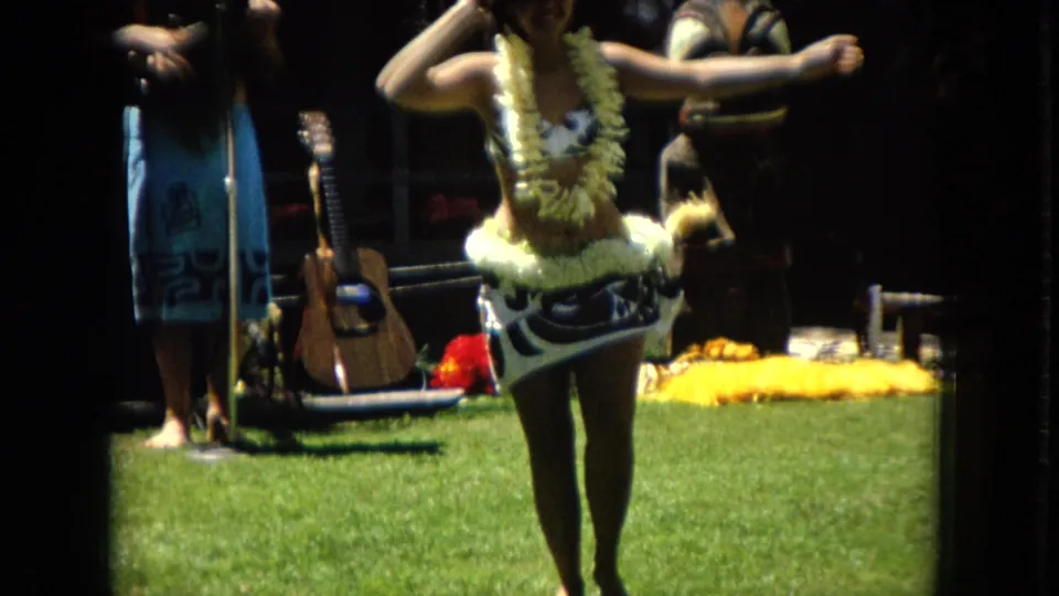 1967 Hawaii home-movie frame showing a hula dancer with raised arms and floral costume details, with instruments and musicians behind her on grass.