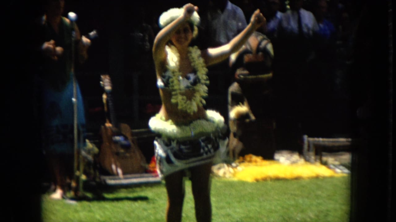In 1967 Hawaii Super 8 footage, a hula dancer wearing leis and a floral headpiece performs on a grassy outdoor area while musicians stand behind her in shade.