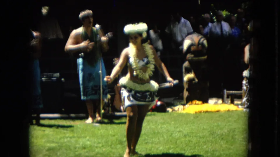 A woman in hula attire steps forward across a grassy performance area in 1967 Hawaii footage, with a musician and other figures visible in the shaded background.