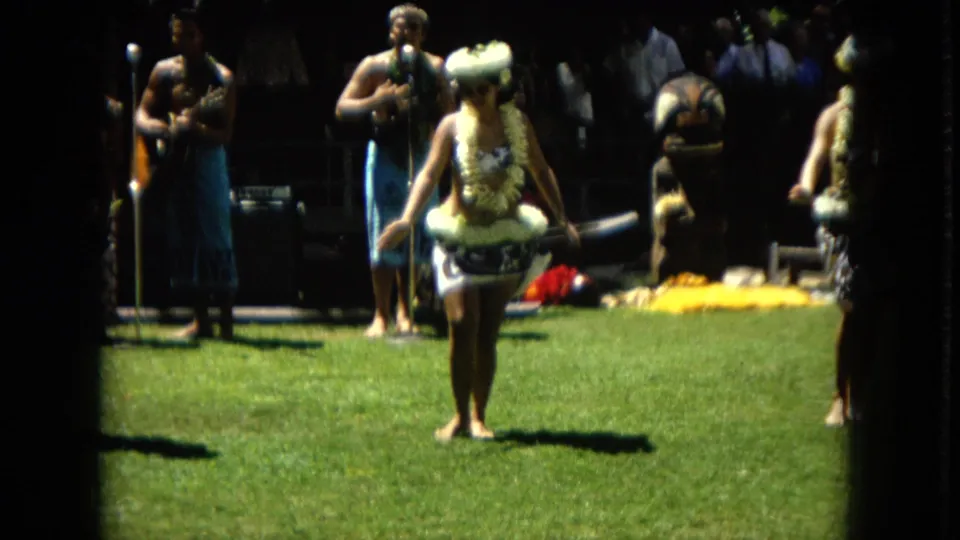 A hula dancer stands centered in the frame during an outdoor 1967 Hawaii performance, with musicians, microphone stands, and another partially visible performer around her.