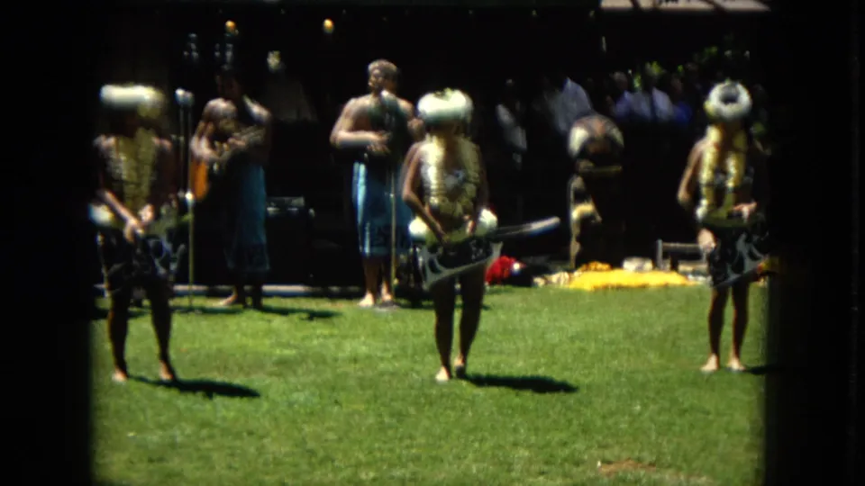 Several hula performers and musicians appear together on a grassy outdoor stage area in 1967 Hawaii Super 8 footage, with the band set back in shade.