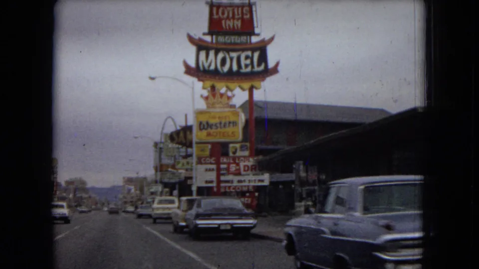 1967 Las Vegas street view from a moving vehicle, with large motel signs, mid-century cars, and commercial storefronts along a broad road.