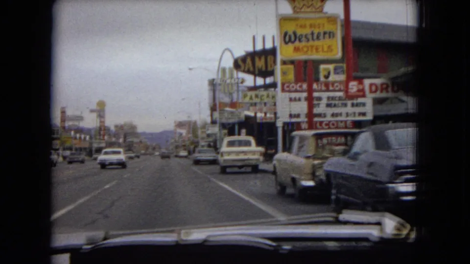 Vehicle-mounted 1967 Las Vegas street view showing traffic, roadside motel signage, a drugstore sign, and distant hills beyond the commercial strip.