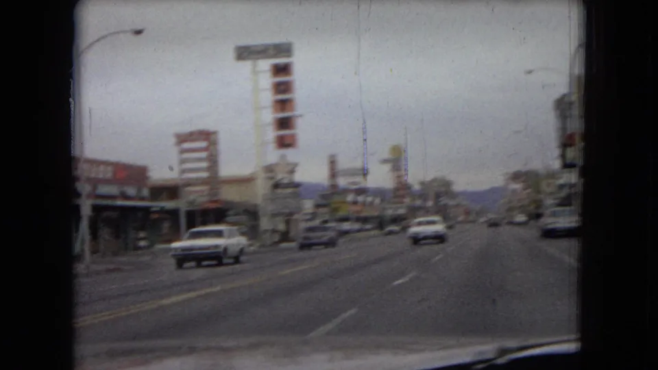 Wide 1967 Las Vegas roadway view with moving cars, low commercial buildings, roadside signs, and muted mountains beyond the street.