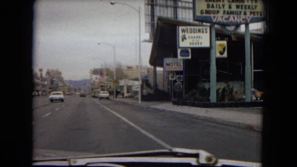 1967 Las Vegas street view from a moving car beside a motel facade with visible vacancy and chapel-wedding signage along the sidewalk.