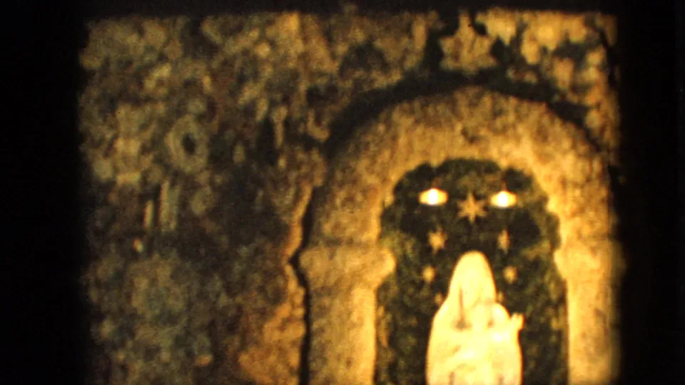 1967 Iowa home-movie frame showing the right side of a grotto-style arched alcove with a white Virgin Mary and child statue partly visible below warm lights and star shapes.