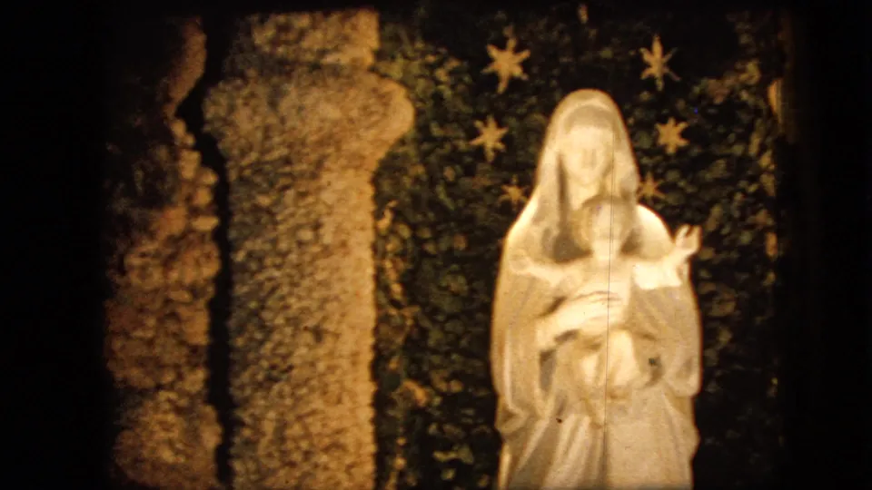 Close 1967 Iowa 8mm view of the white Virgin Mary and infant Jesus statue, with rough stonework on one side and a dark backdrop decorated with star shapes behind it.