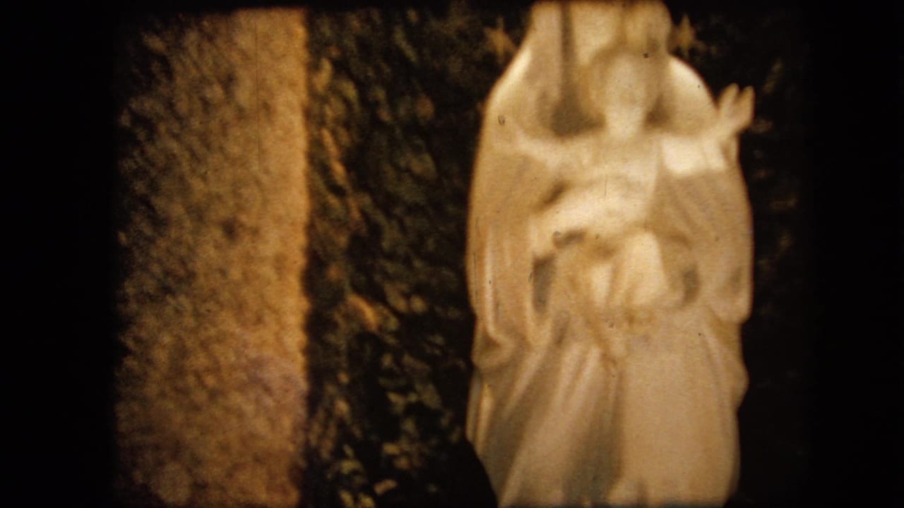 1967 Iowa 8mm frame showing a white Virgin Mary and infant Jesus statue lit warmly against a dark grotto-like or foliage-backed setting.