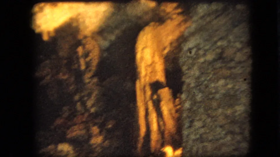 1967 Iowa 8mm frame showing a warm illuminated grotto-like surface with a small dark cross-like form and surrounding rough texture.