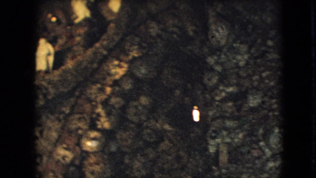 1967 Iowa 8mm frame showing a dark rocky hillside or stone ruin area, with small distant visitors partly visible near the upper left and a bright vertical mark near the center-right.