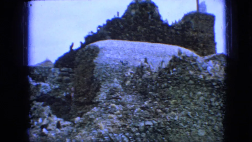 1967 Iowa 8mm frame showing rocky stone ruins, a pale wall or path surface, greenery, and small distant visitors near the lower left edge.