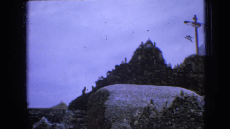 1967 Iowa 8mm frame showing a rocky mound above a pale wall or path, with cloudy sky and a slender post or sign structure on the right.