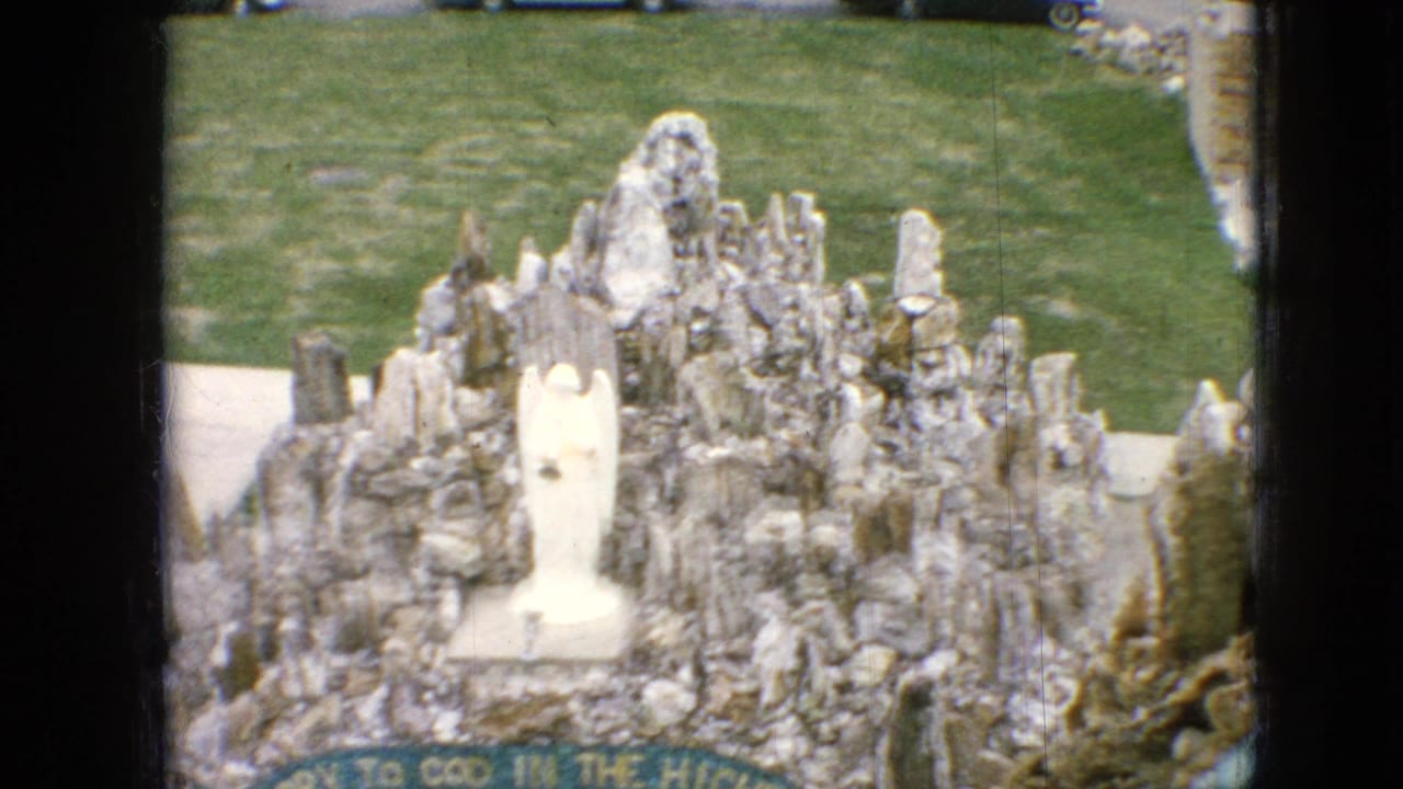 1967 Iowa 8mm frame showing a rocky outdoor grotto with a central white statue, grass behind it, and a green banner along the lower edge.