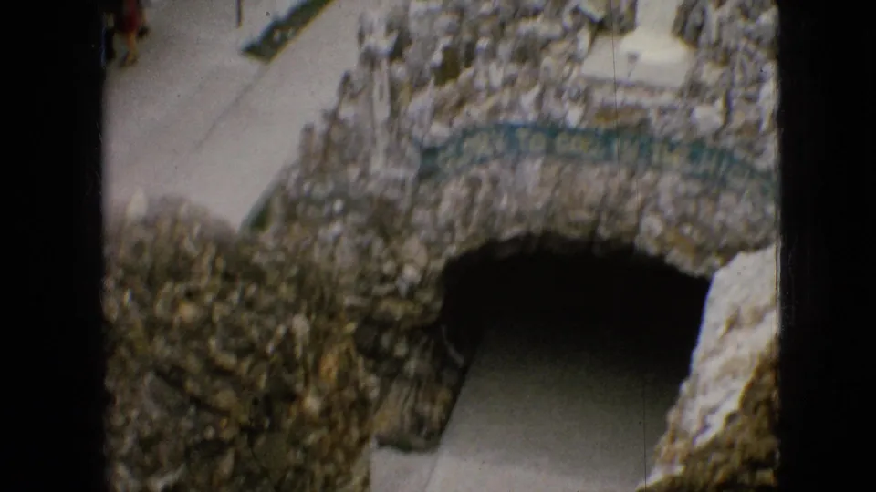 1967 Iowa 8mm frame showing the grotto's stone arch and dark passageway, with sidewalk, low stone walls, and the green religious banner above the opening.