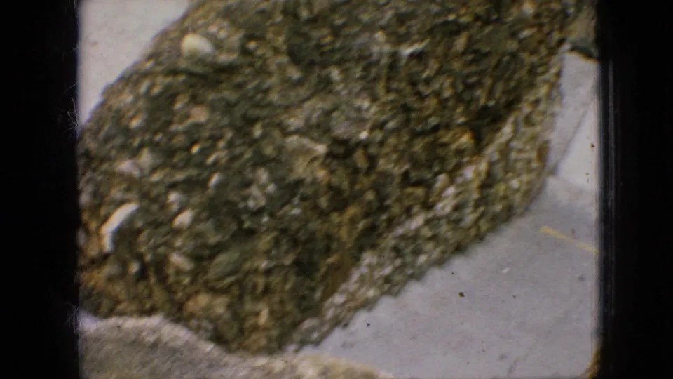 1967 Iowa home-movie frame showing a close view of the grotto's rough stone wall, with a paved sidewalk and the edge of the surrounding outdoor setting.