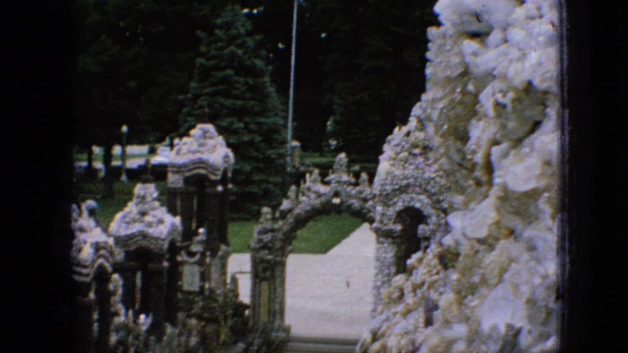 1967 Iowa 8mm frame showing a shell- and stone-covered outdoor passage with arched openings, steps, lawn, trees, and dark film borders.