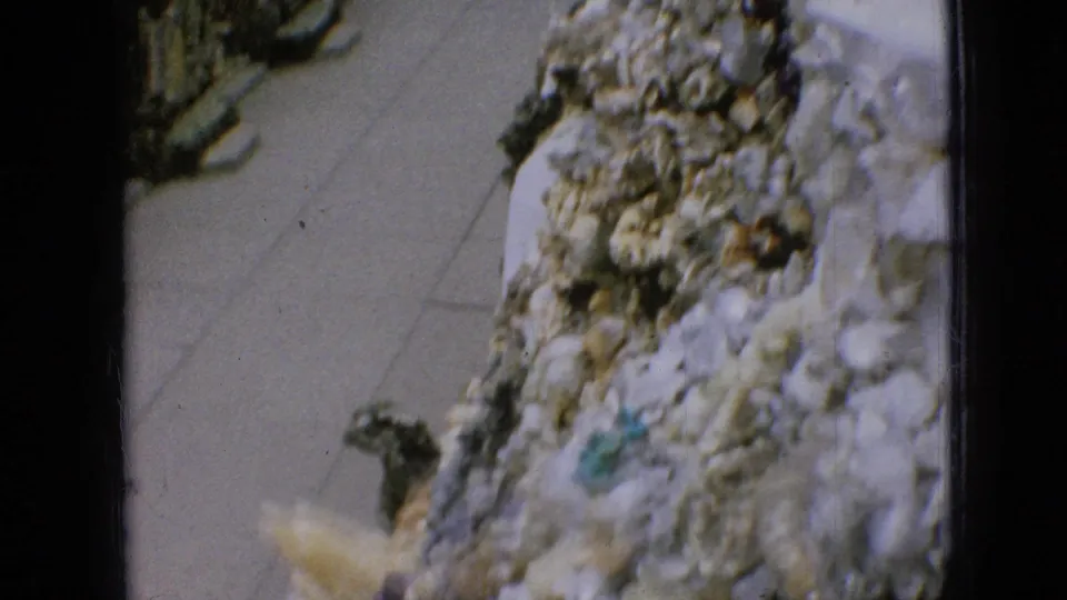 Close 1967 Iowa 8mm view of a textured shell-and-stone wall beside a paved walkway, with part of an arch and film edge visible.