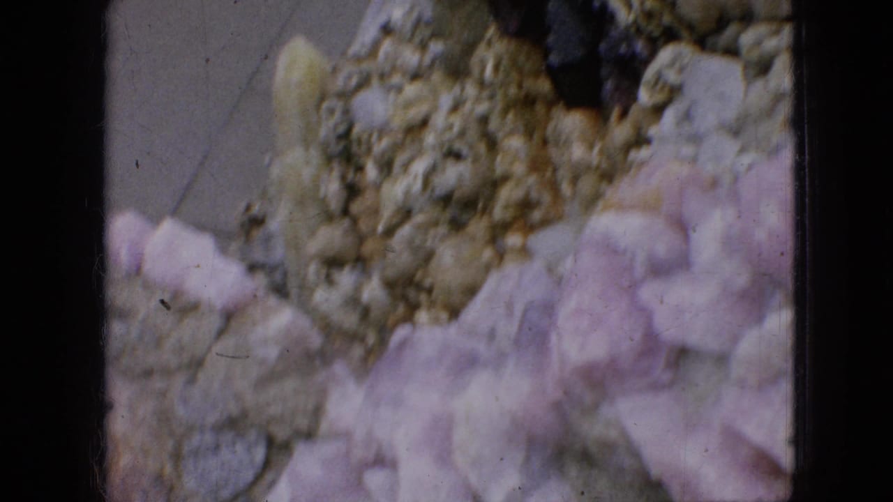 1967 Iowa 8mm frame showing a close, soft-focused view of rough gravel-coated masonry with pale pink and white stone fragments in the foreground.