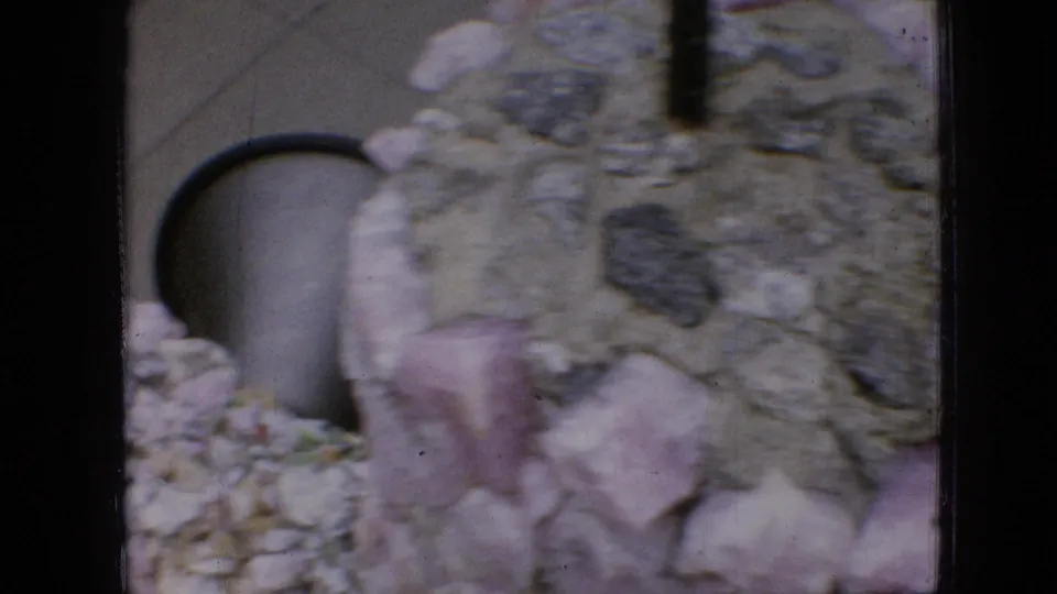 1967 Iowa home-movie frame showing a textured gravel wall, pale stone pieces, and a dark arched opening partly obscured by the close camera angle.