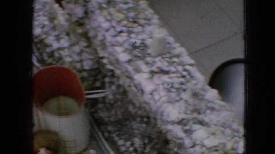 1967 Iowa 8mm frame showing a curving rough stone wall, metal railing, a dark arched opening, and a red-topped object near the lower left.