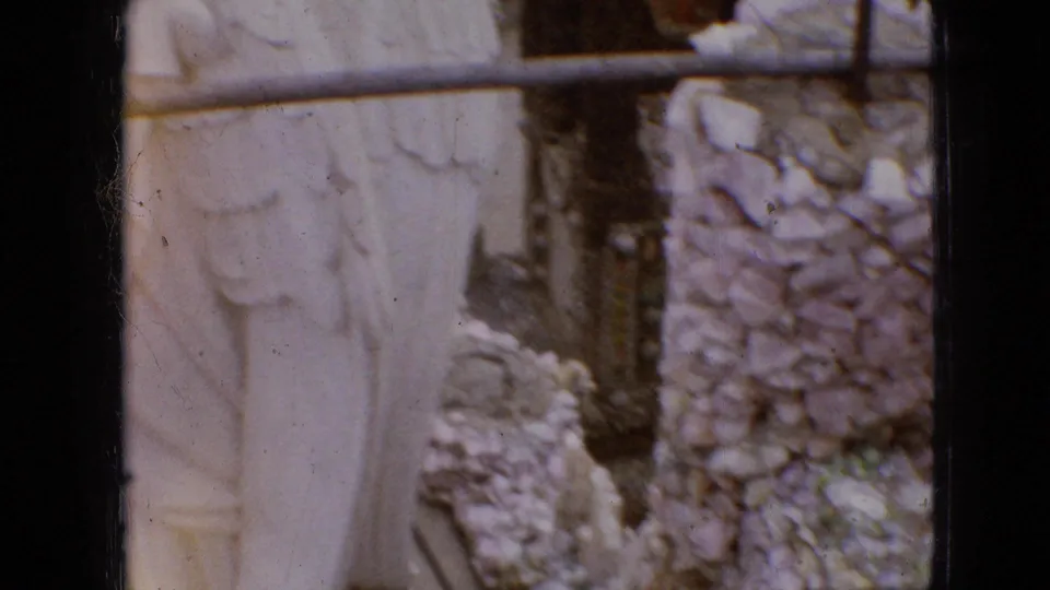 1967 Iowa home-movie frame showing a pale angel statue partly hidden by rough stonework, carved panels, debris, and a horizontal metal rail.