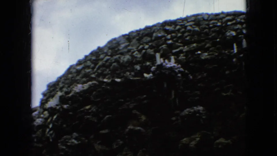 1967 Iowa 8mm frame looking past dark stone edges toward a rounded mass of rough stacked stone beneath an overcast sky.