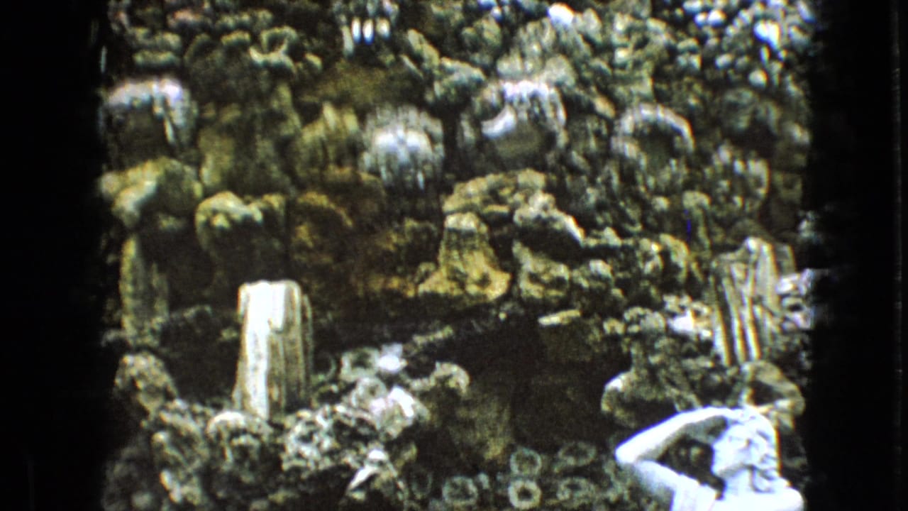 1967 Iowa 8mm frame showing a white classical statue partly cropped at the lower right, set against a textured rock grotto with layered stone and small upright forms.