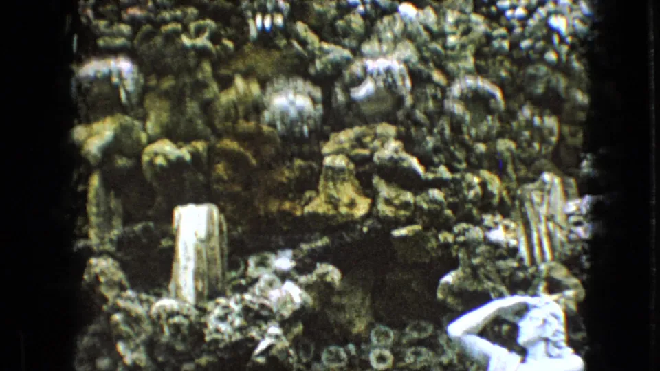 1967 Iowa 8mm frame showing a white classical statue partly cropped at the lower right, set against a textured rock grotto with layered stone and small upright forms.
