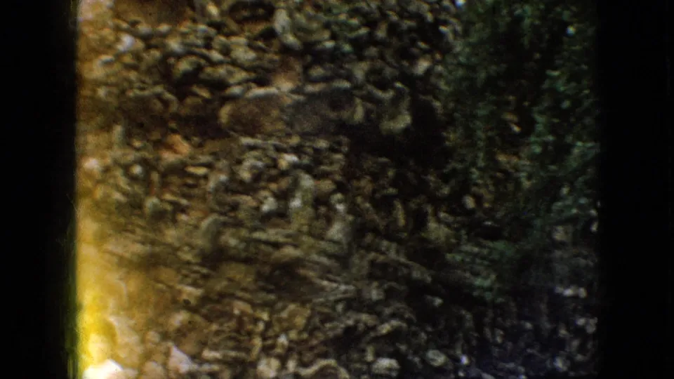 1967 Iowa 8mm frame focused on the rough layered rock surface of the grotto, with dark shadowed areas and a warm yellow flare along the lower left edge.
