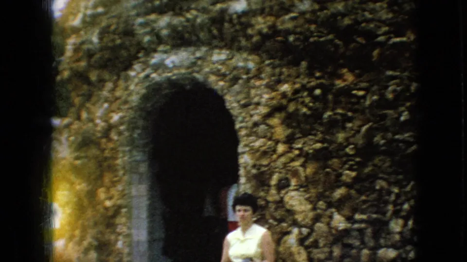1967 Iowa home-movie frame showing a woman in a light-colored dress standing near a dark arched stone entrance within the textured rock grotto wall.