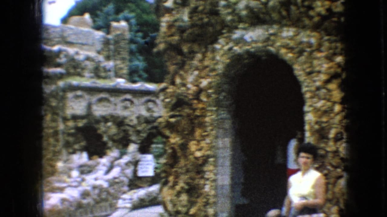 1967 Iowa home-movie frame showing an adult visitor near a shadowed stone archway, with rough masonry, small arched alcoves, and trees around the garden structure.