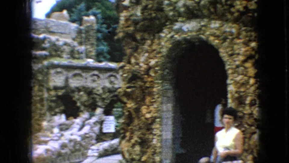 1967 Iowa home-movie frame showing an adult visitor near a shadowed stone archway, with rough masonry, small arched alcoves, and trees around the garden structure.
