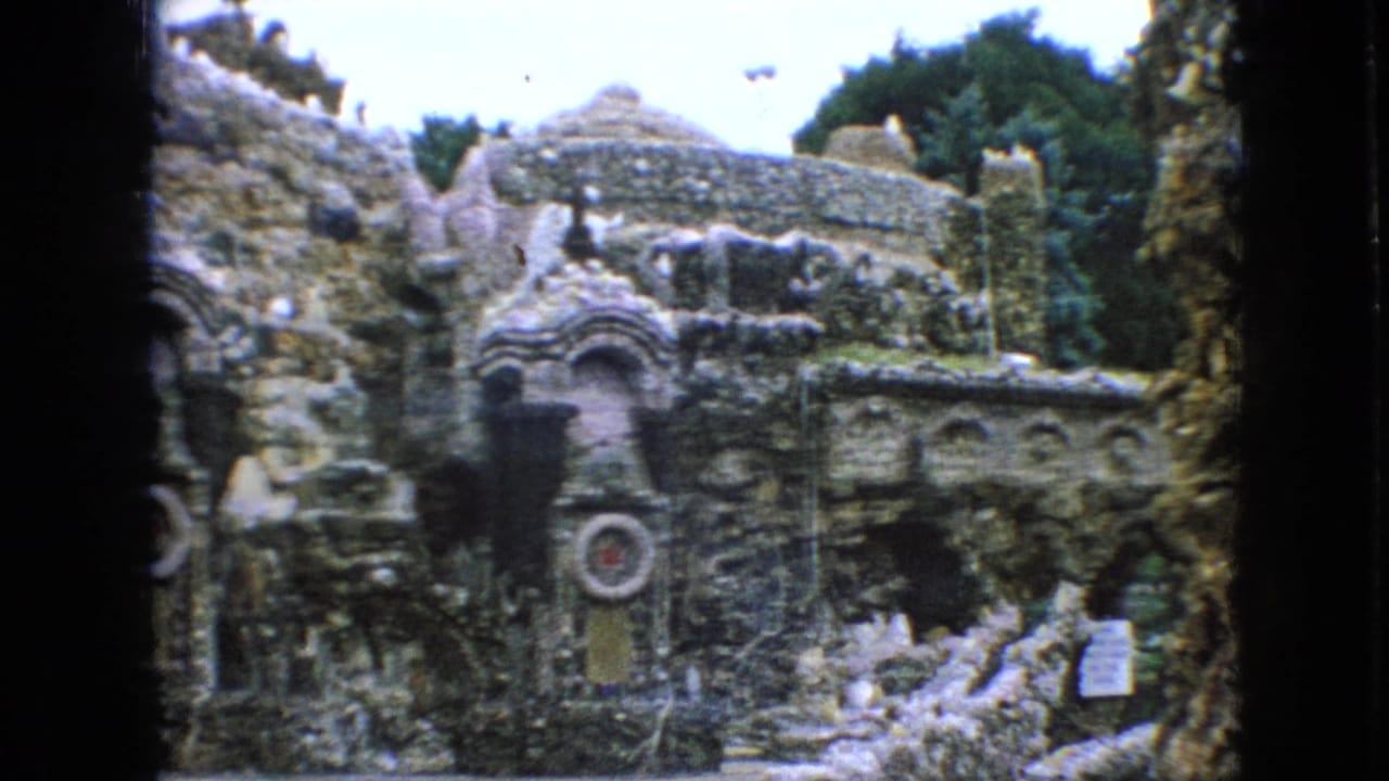 1967 Iowa 8mm frame centered on an ornate rock garden facade with layered stonework, arched niches, decorative forms, posted signs, and trees above the structure.