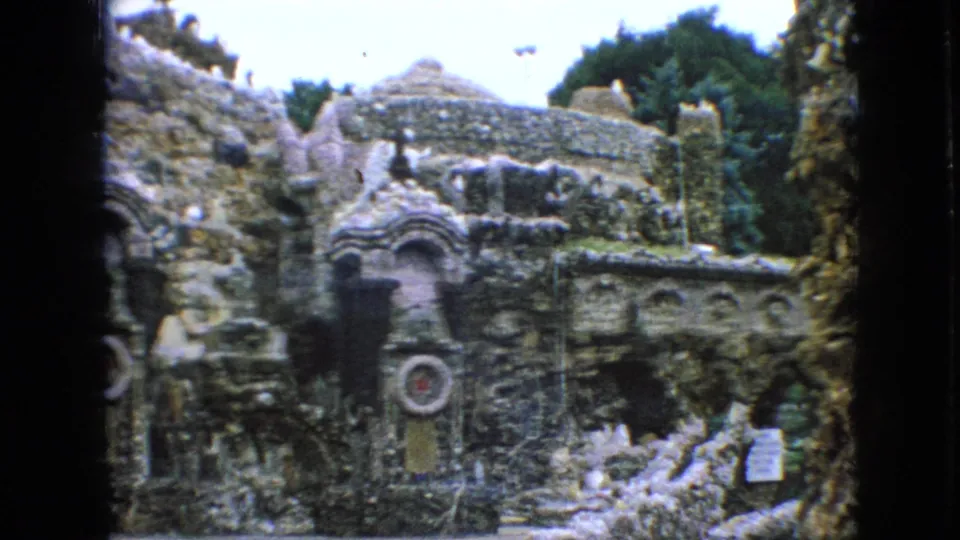 1967 Iowa 8mm frame centered on an ornate rock garden facade with layered stonework, arched niches, decorative forms, posted signs, and trees above the structure.