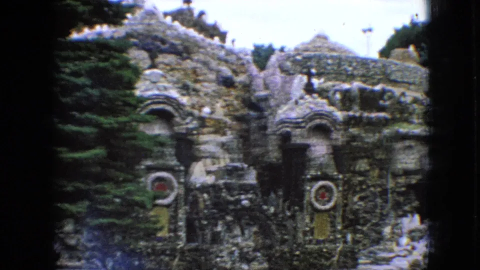 1967 Iowa home-movie view of a grotto-like stone garden structure, showing textured rockwork, arched alcoves, small signs, dark openings, and greenery around the site.