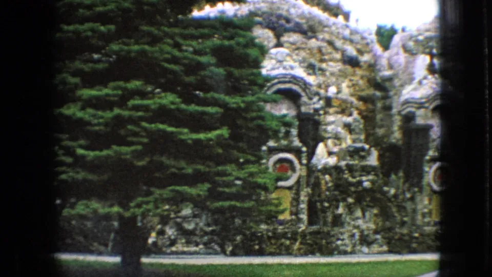1967 Iowa 8mm frame showing a wider view of the stone garden, with arched masonry, dense green trees, a small lawn, and a partially visible adult visitor near the right side.