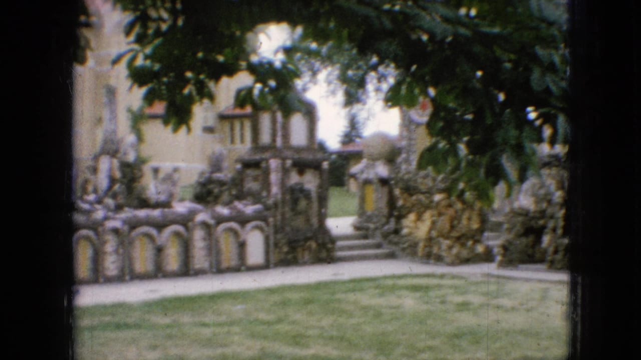 1967 Iowa home-movie frame with a pale building behind ornate stone garden walls, rounded alcoves, walkway steps, lawn, and dark tree foliage at the top edge.