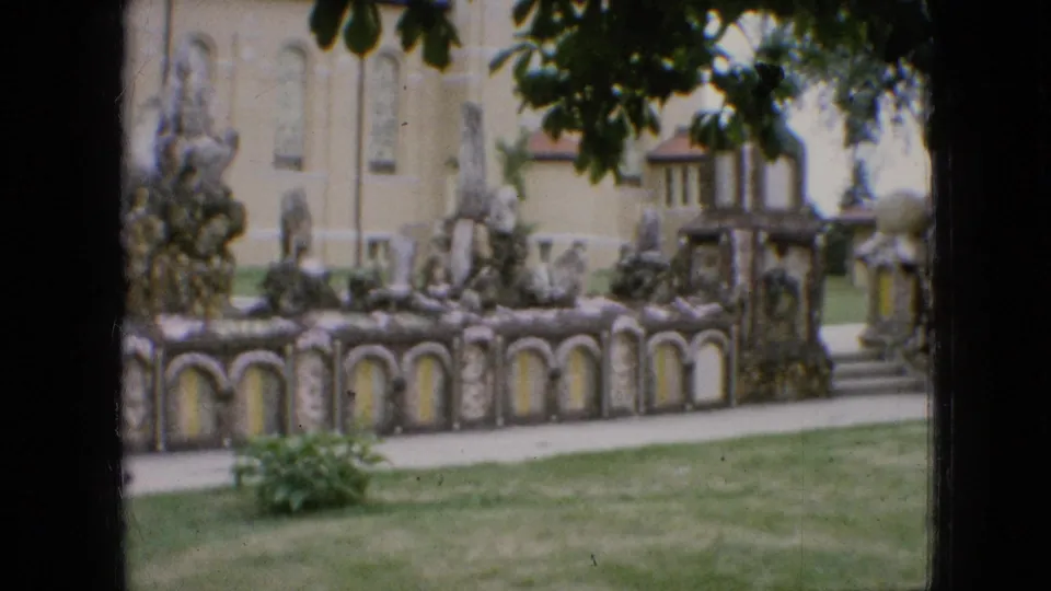 1967 Iowa 8mm frame showing a closer view of arched stone garden walls, irregular rockwork, yellow panels, a pale building facade, lawn, and foreground foliage.