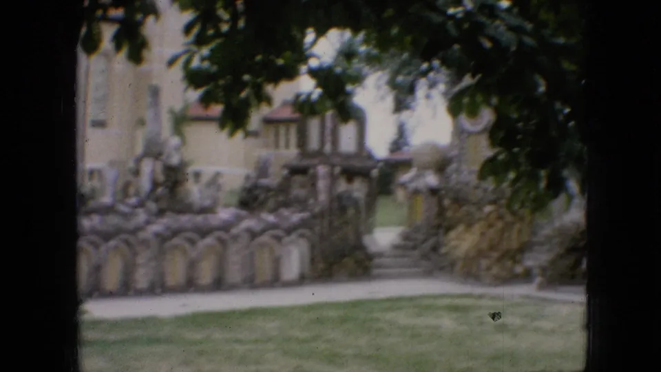 1967 Iowa home-movie frame showing a stone garden entrance or passage with steps, arched wall details, lawn, pale building background, and heavy tree cover overhead.