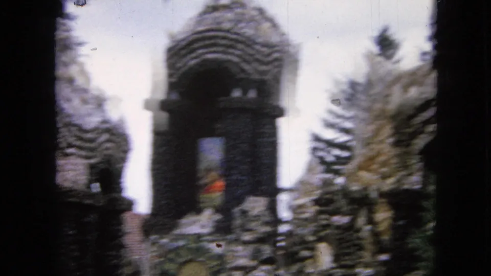 1967 Iowa 8mm frame showing a soft-focus stone tower with an arched opening, surrounding sculptural walls, and trees against a bright sky.