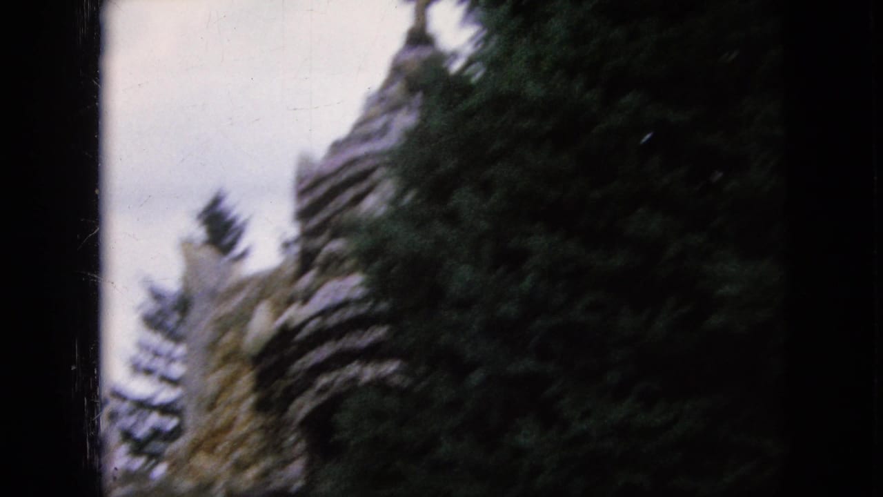 1967 Iowa 8mm frame showing a blurred older stone structure partly hidden by dark conifer trees under an overcast sky.