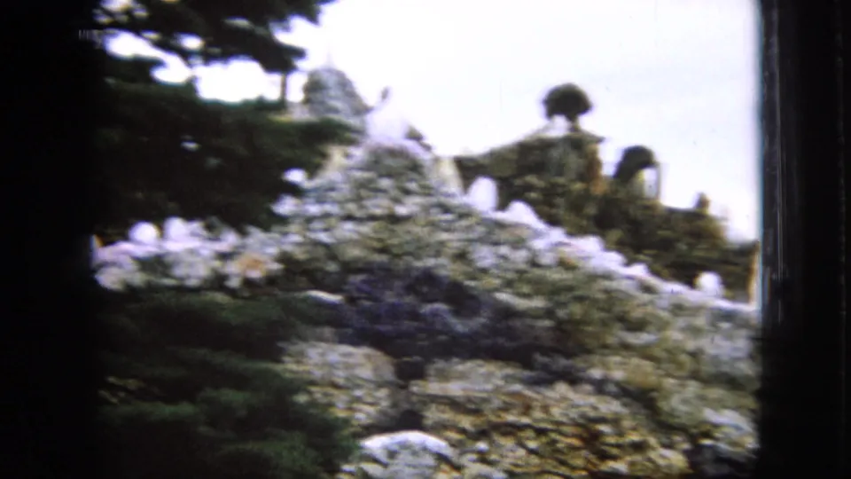 1967 Iowa 8mm frame showing a stone structure topped with Christian crosses, viewed from below through dark conifers and bright sky.
