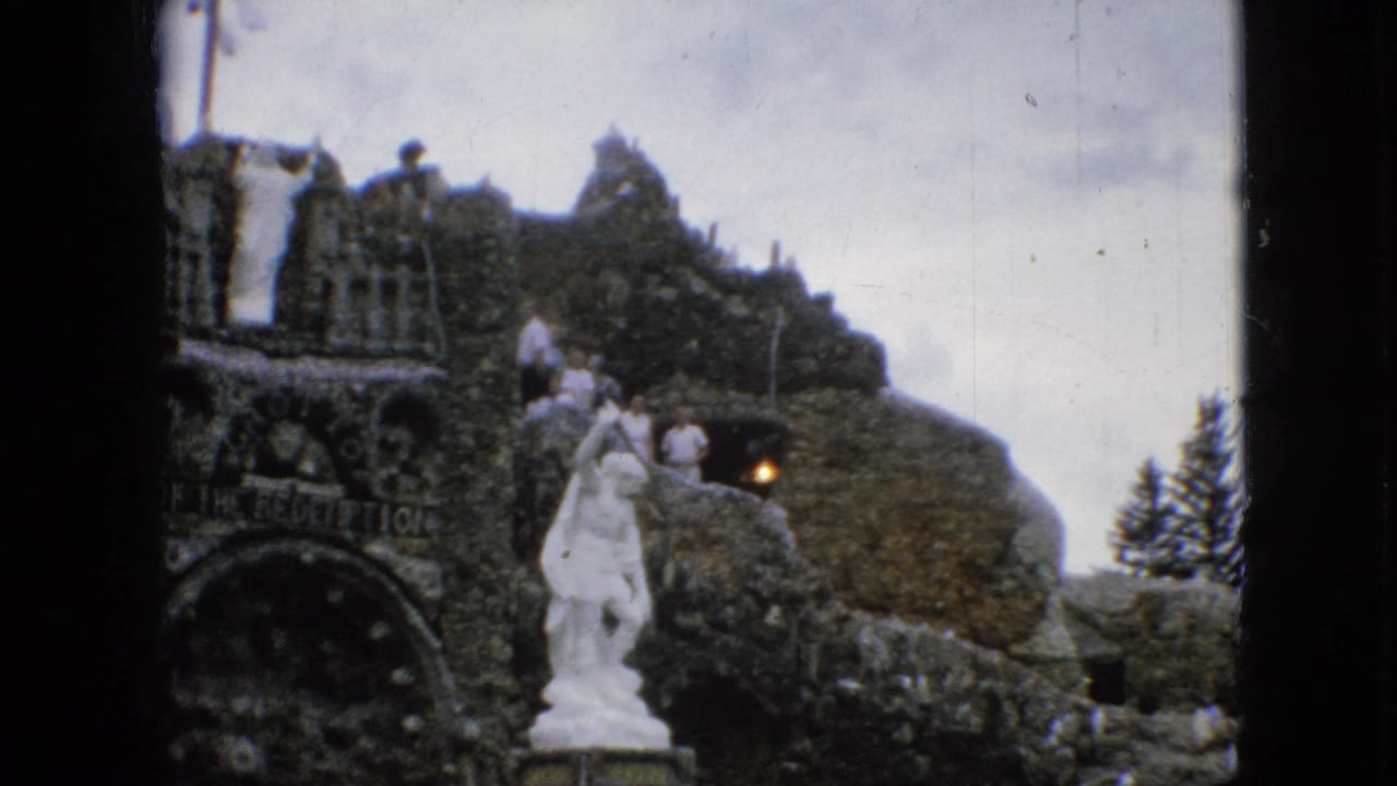 1967 Iowa vacation frame showing visitors on the elevated stonework of the Grotto of the Redemption, with a white statue in front.