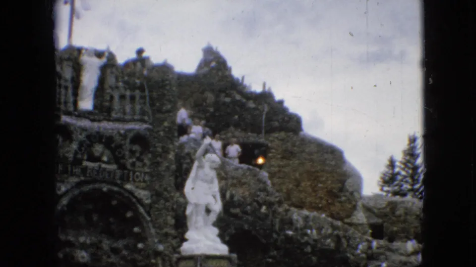 1967 Iowa vacation frame showing visitors on the elevated stonework of the Grotto of the Redemption, with a white statue in front.