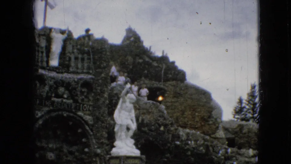 1967 Iowa 8mm frame of the Grotto of the Redemption with a white statue, rough stonework, and distant visitors on the upper level.