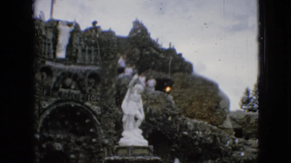 1967 Iowa home-movie frame showing the Grotto of the Redemption stonework, white statue, distant visitors, and visible 8mm contact-sheet borders.
