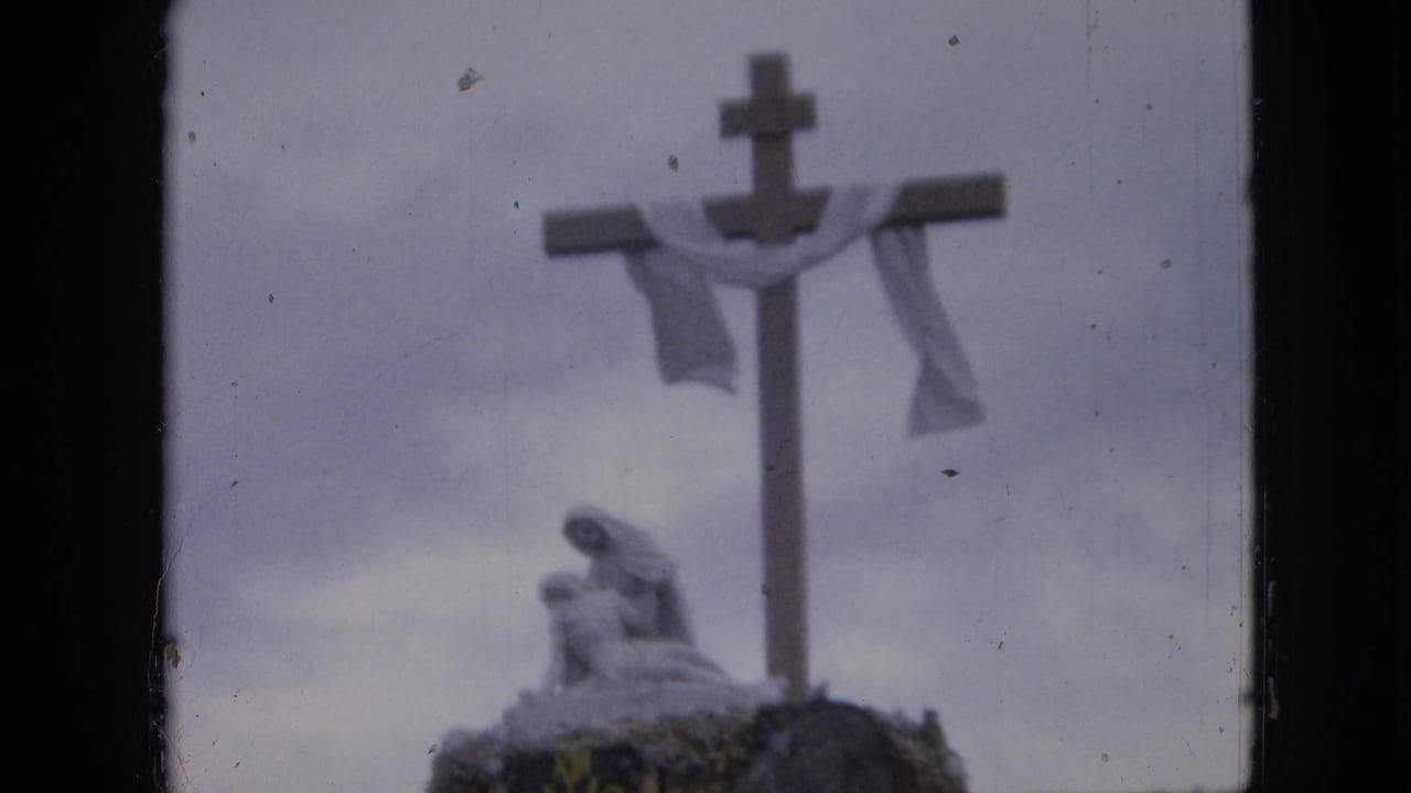 1967 Iowa 8mm frame showing a large outdoor cross with a white drape and a statue-like figure near its base, seen in soft focus.
