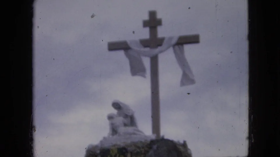 1967 Iowa 8mm frame showing a large outdoor cross with a white drape and a statue-like figure near its base, seen in soft focus.