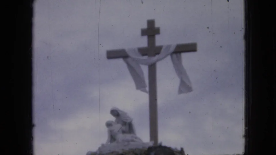1967 Iowa home-movie frame of a religious cross draped with cloth, with a statue-like figure below and visible 8mm frame borders.