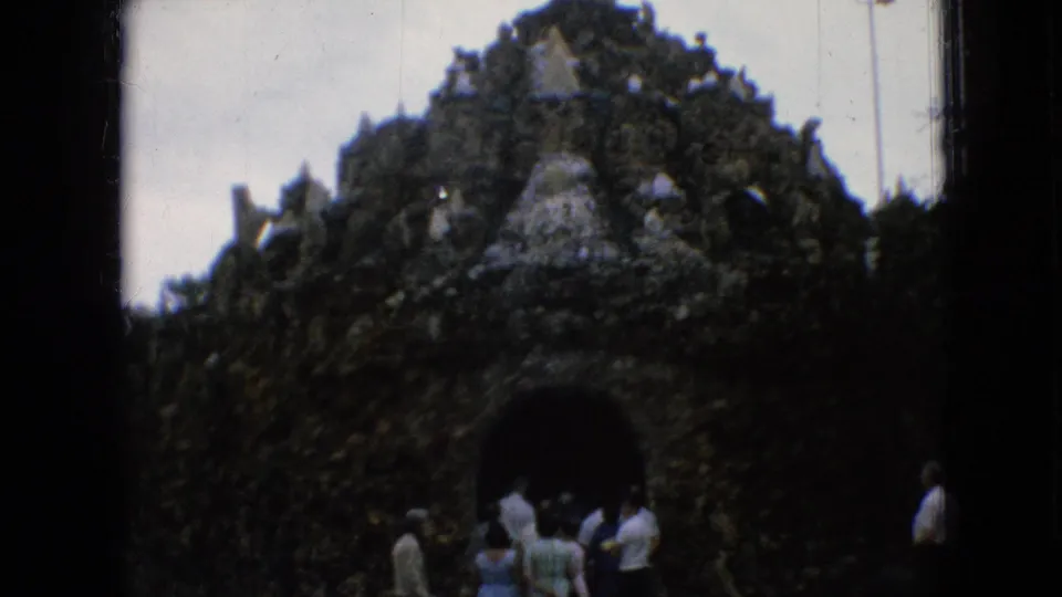 1967 Iowa 8mm frame showing several visitors outside a large stone or grotto-like shrine structure with an arched tunnel entrance.